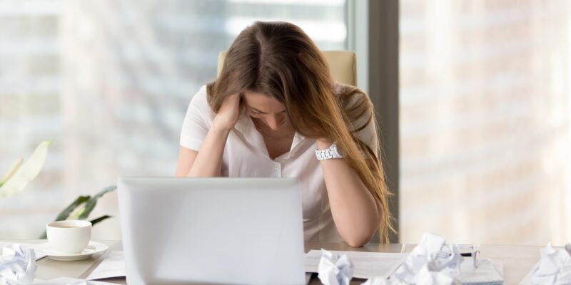 Desperate businesswoman sitting at the desk with laptop and crumpled paper. Female office worker suffers because of too much mess in her thoughts. Stressed woman entrepreneur feeling creativity crisis